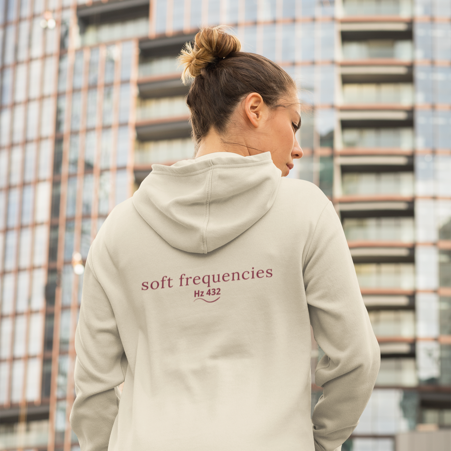 Woman wearing Soft Frequencies™ hoodie in Solace with “soft frequencies Hz 432” text on back, standing in front of modern glass building
